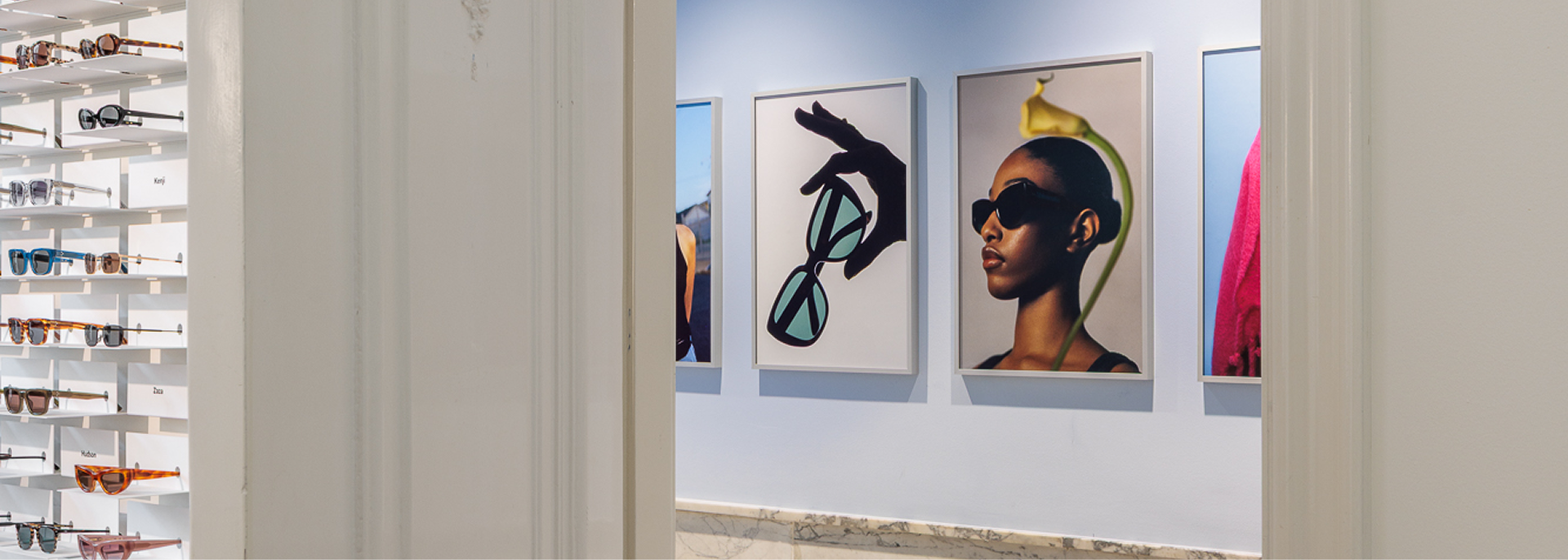 A well-lit hallway inside the Ace & Tate headquarters with product displays on the left and large, framed artwork of models wearing sunglasses on the right wall.